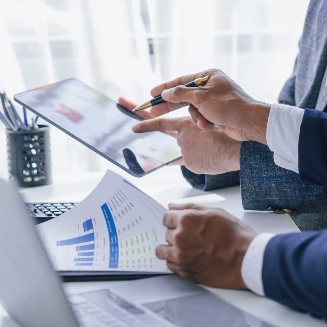 A man's hand holds a pen pointing at coworker's tablet while sitting on a desk with other documents displaying financial data.